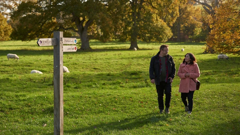 Two visitors walking between sheep with a finger sign pointing in three directions in the parkland at Montacute House